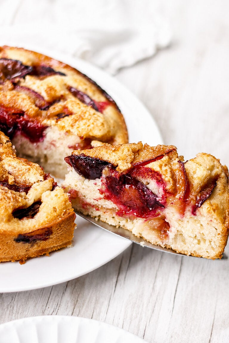 A slice of Easy Italian Plum Cake is being lifted from a larger round cake on a white plate. The cake has a golden crust and visible baked plum slices throughout.