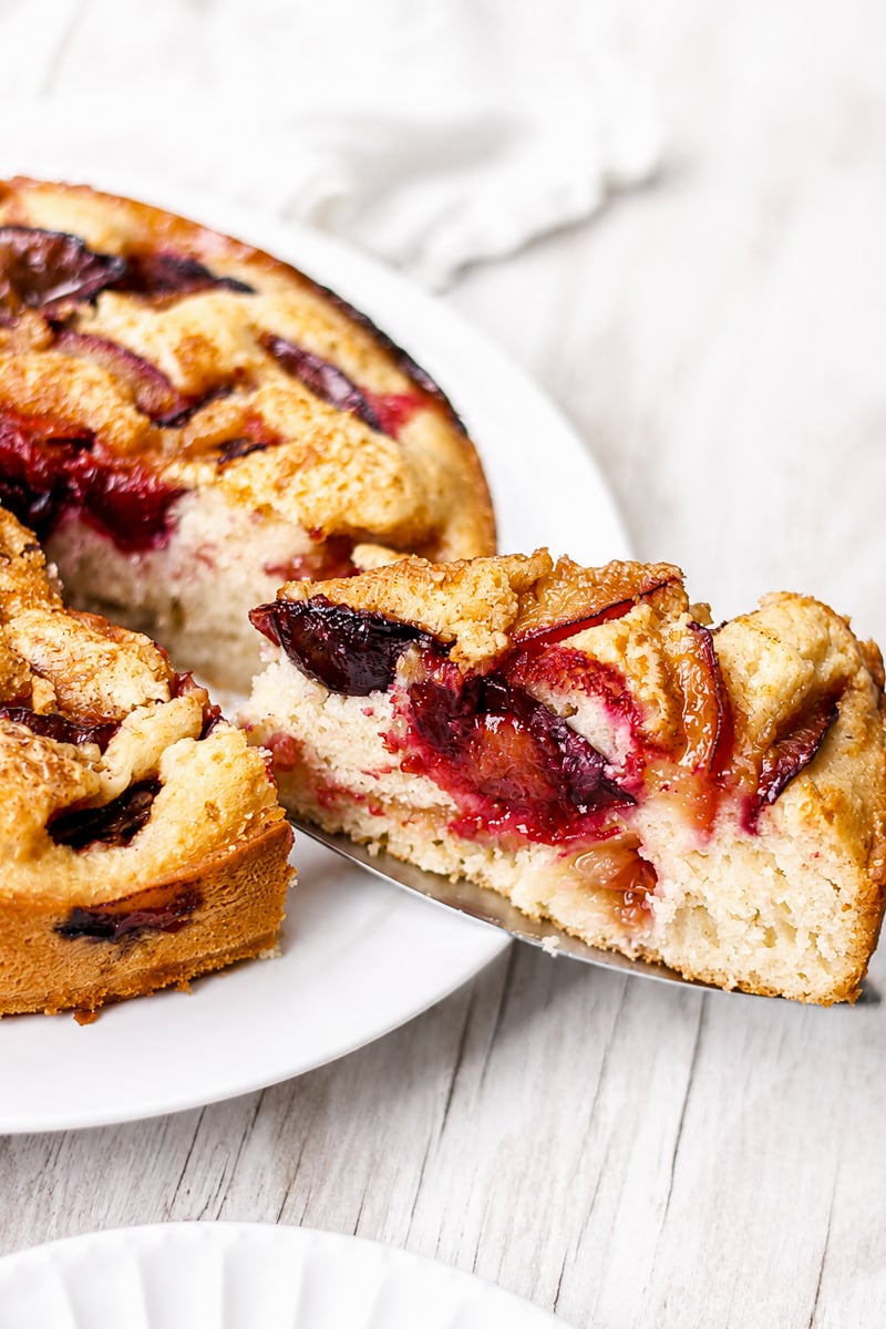 A slice of Easy Italian Plum Cake is being lifted from a larger round cake on a white plate. The cake has a golden crust and visible baked plum slices throughout.
