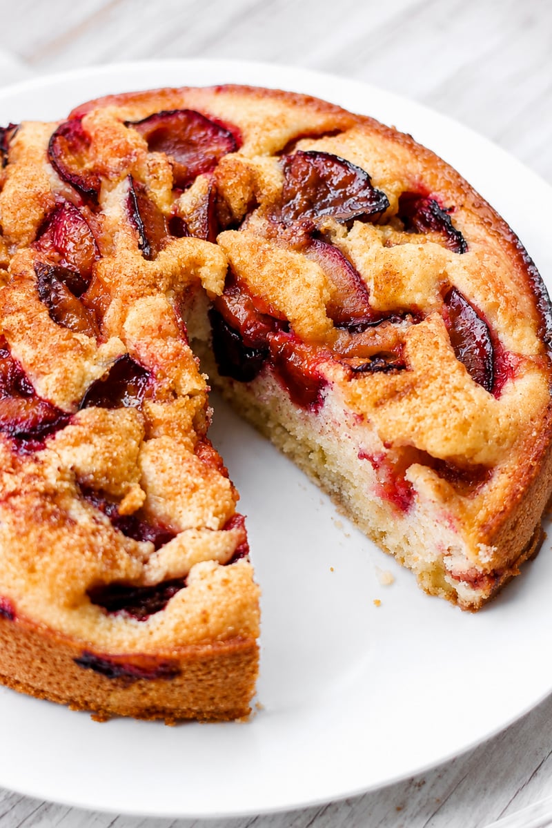 A round Easy Italian Plum Cake with a golden crust sits on a white plate. One slice is missing, revealing the moist, light interior topped with baked plum slices.
