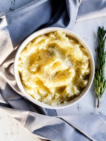 A bowl of Instant Pot mashed potatoes topped with melted butter and black pepper, next to a sprig of rosemary on a cloth-covered table.