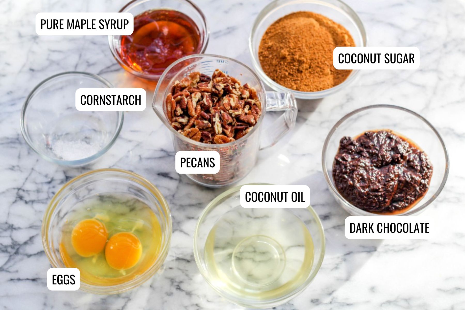 Various baking ingredients for Chocolate Pecan Pie are arranged in bowls on a marble surface, including eggs, coconut oil, pecans, cornstarch, pure maple syrup, coconut sugar, and dark chocolate. Each ingredient is labeled.