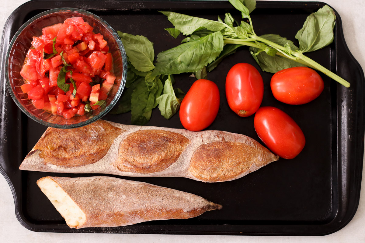 image of bread, chopped tomatoes in a glass dish, basil and more tomateos on a baking sheet.