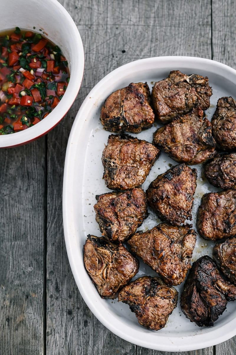 Grilled Lamb Chops with Ammoglio Sauce A white dish filled with Grilled Lamb Chops with Ammoglio Sauce sits on a rustic wooden surface. Next to it, there is a bowl of colorful chimichurri boasting chopped herbs, red onion, and tomato.