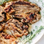 Three grilled lamb chops on a floral-patterned plate, garnished with fresh rosemary sprigs. The meat is browned and slightly charred, perfectly prepared as Grilled Lamb Chops with Ammoglio Sauce for a flavorful finish.