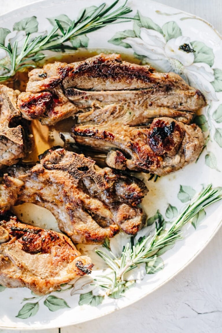 Three grilled lamb chops on a floral-patterned plate, garnished with fresh rosemary sprigs. The meat is browned and slightly charred, perfectly prepared as Grilled Lamb Chops with Ammoglio Sauce for a flavorful finish.