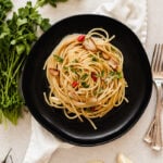 A black plate of spaghetti aglio e olio garnished with parsley, sliced garlic, and red chili peppers rests on a white napkin. Fresh parsley, red chili peppers, garlic cloves, and two forks surround the dish.