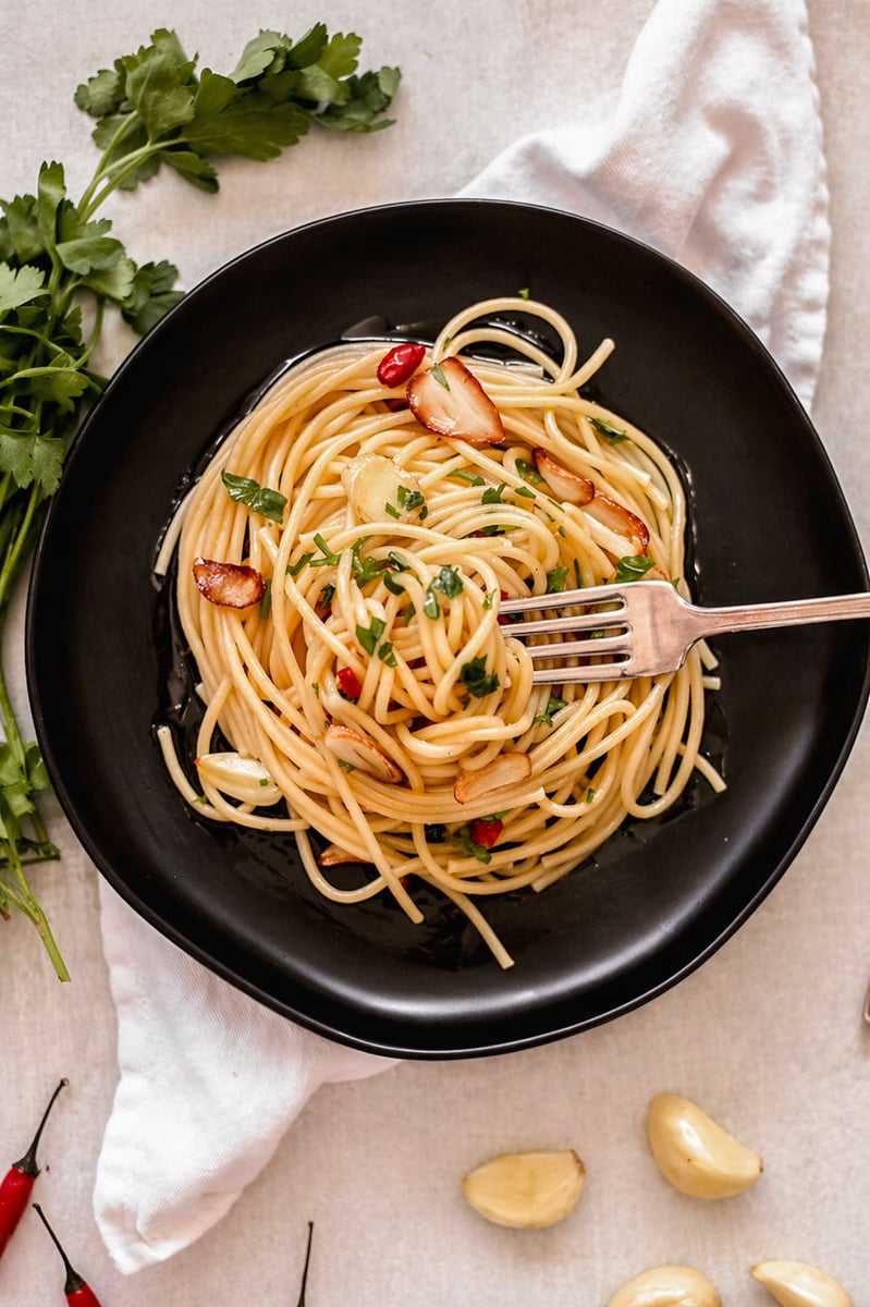 A black plate of spaghetti aglio e olio is topped with sliced garlic, chopped parsley, and red chili slices. A fork rests on the pasta. Fresh parsley, chili peppers, and garlic cloves are nearby on a white cloth background.
