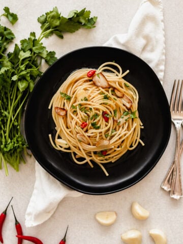A black plate of spaghetti aglio e olio garnished with parsley, sliced garlic, and red chili peppers rests on a white napkin. Fresh parsley, red chili peppers, garlic cloves, and two forks surround the dish.