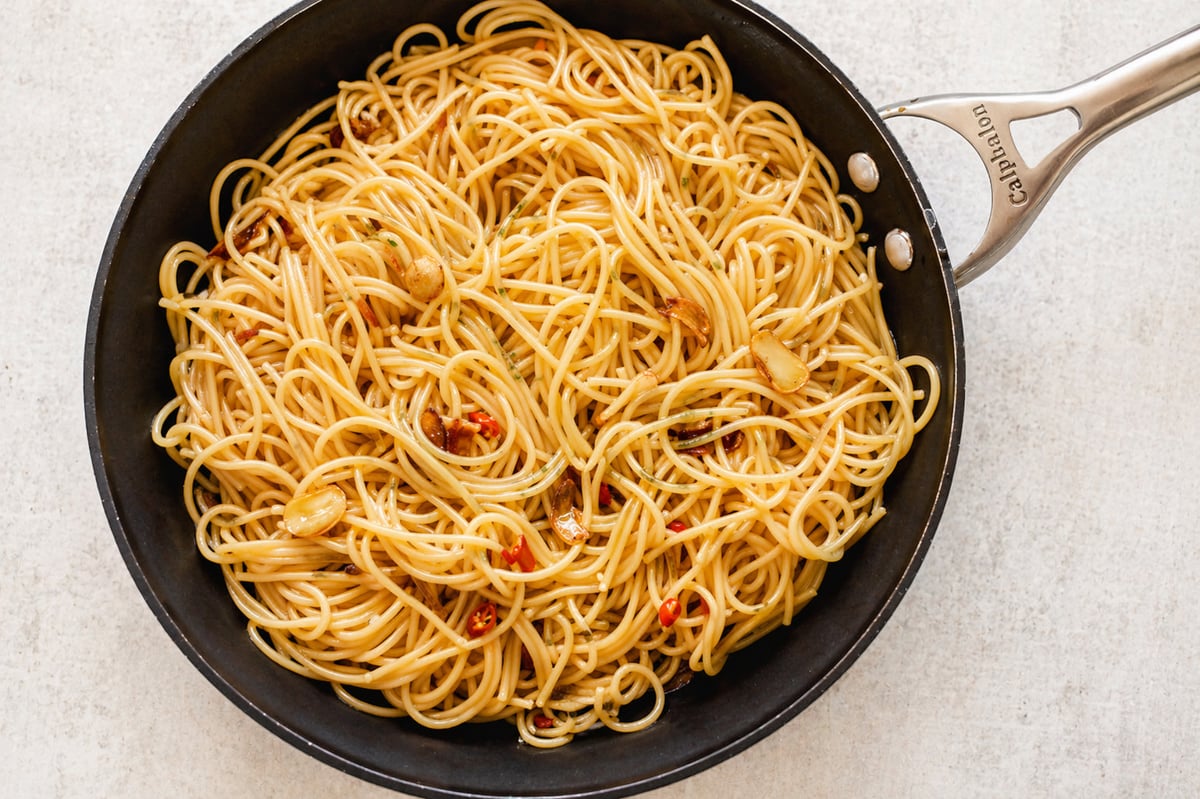 A black frying pan filled with classic spaghetti aglio e olio, garnished with slices of garlic and red chili flakes, resting on a light-colored surface.