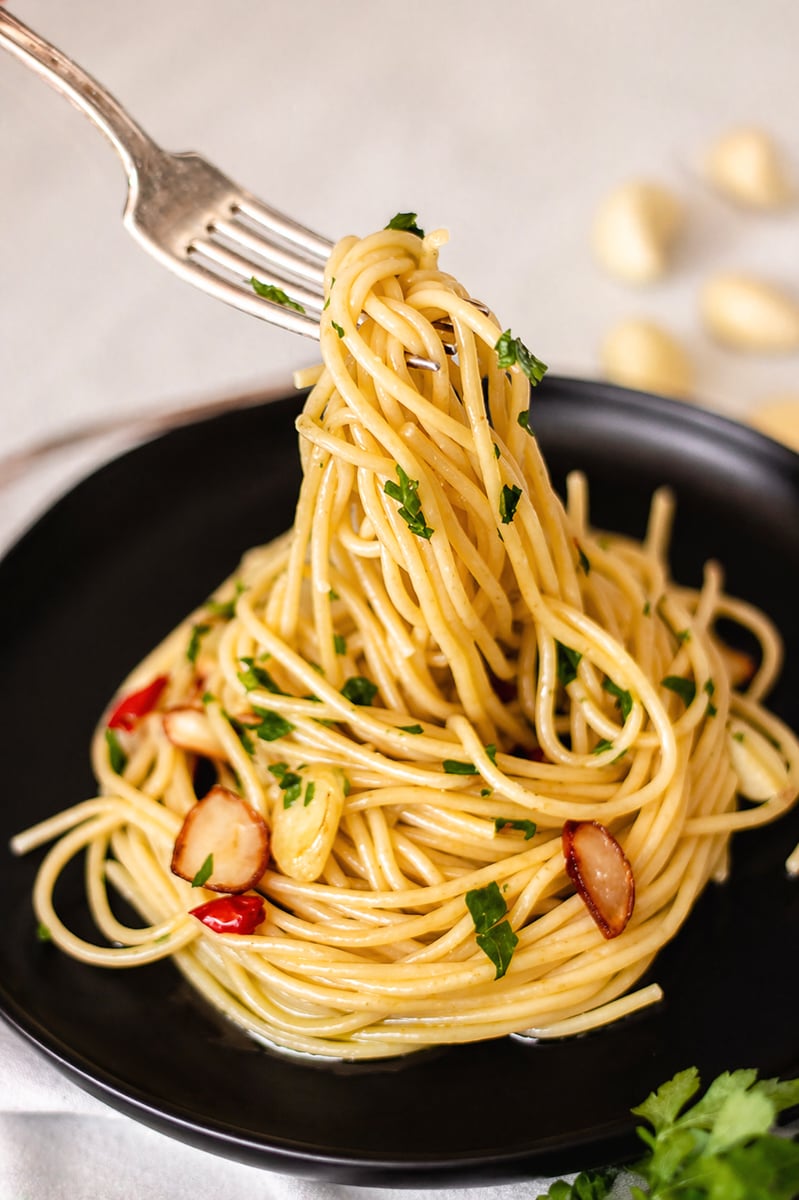 A fork twirls spaghetti aglio e olio on a black plate, garnished with chopped parsley, thinly sliced garlic, and red chili. Garlic cloves and herbs are blurred in the background.