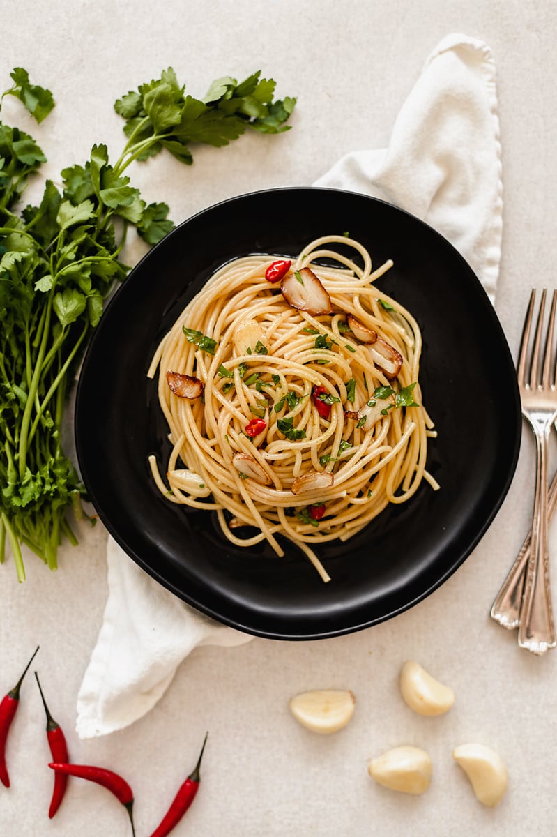 A black plate of spaghetti aglio e olio garnished with parsley, sliced garlic, and red chili peppers rests on a white napkin. Fresh parsley, red chili peppers, garlic cloves, and two forks surround the dish.