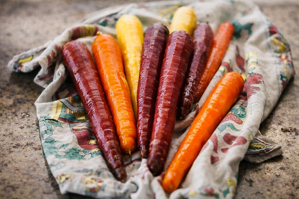 Several colorful carrots-orange, purple, and yellow-are arranged on a patterned cloth atop a stone-like surface. Fresh and vibrant, these carrots vary in size and display their natural texture.