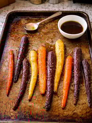 A baking sheet with roasted carrots in vibrant colors arranged in a row, accompanied by a spoon and a small white bowl of dark sauce nestled in the corner of the tray.