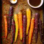 Roasted purple, yellow, and orange carrots sprinkled with sesame seeds are arranged on a baking tray. A silver spoon rests nearby, alongside a small bowl of carrot-infused brown sauce in the top right corner.