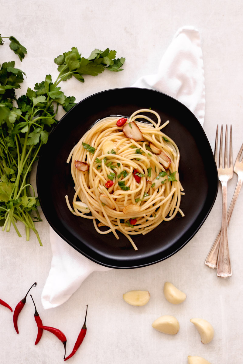 overhead image of pasta on a black plate, forks, parsley, white napkin, hot pepper, garlic in background.