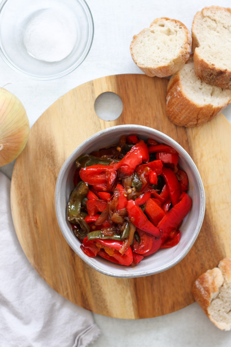 image of red and green peppers in a bowl on a wooden board with bread and onion in background.