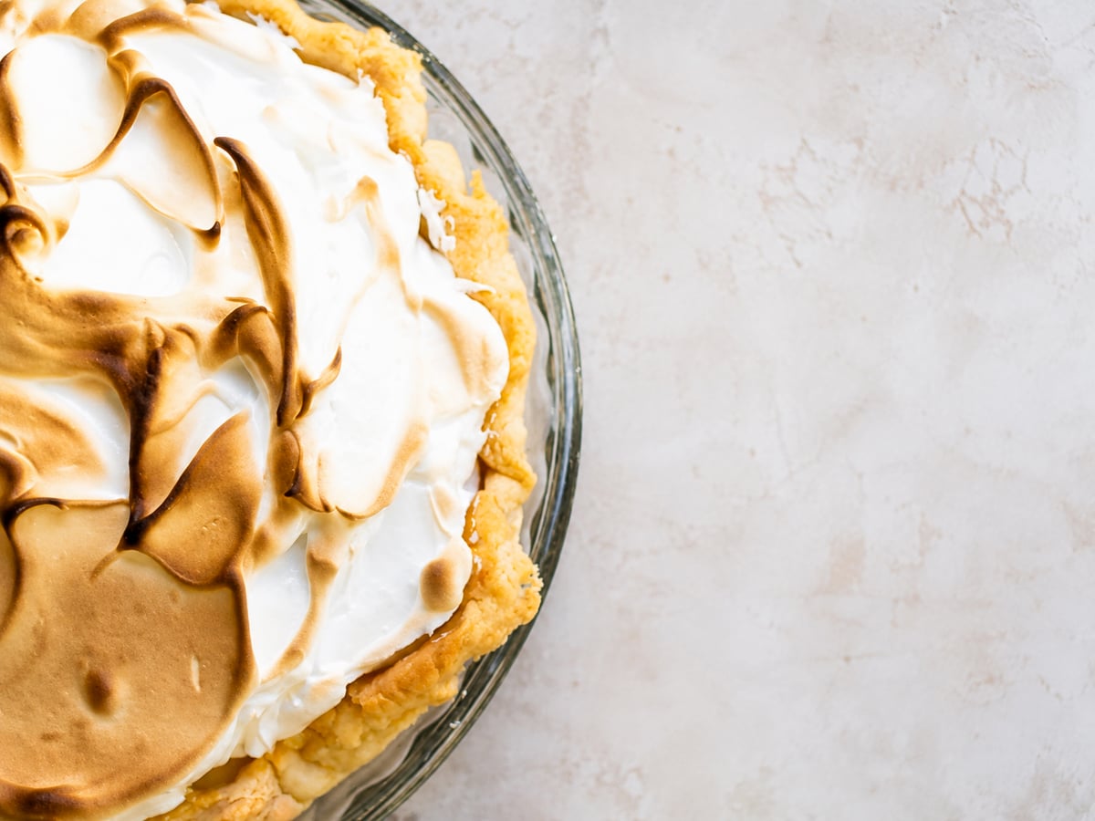 A close-up of a Lemon Meringue Pie with golden-brown toasted meringue peaks in a glass pie dish, set on a light textured surface.