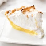 A slice of Lemon Meringue Pie with a golden-brown meringue topping sits on a white plate, with a lemon blurred in the background.
