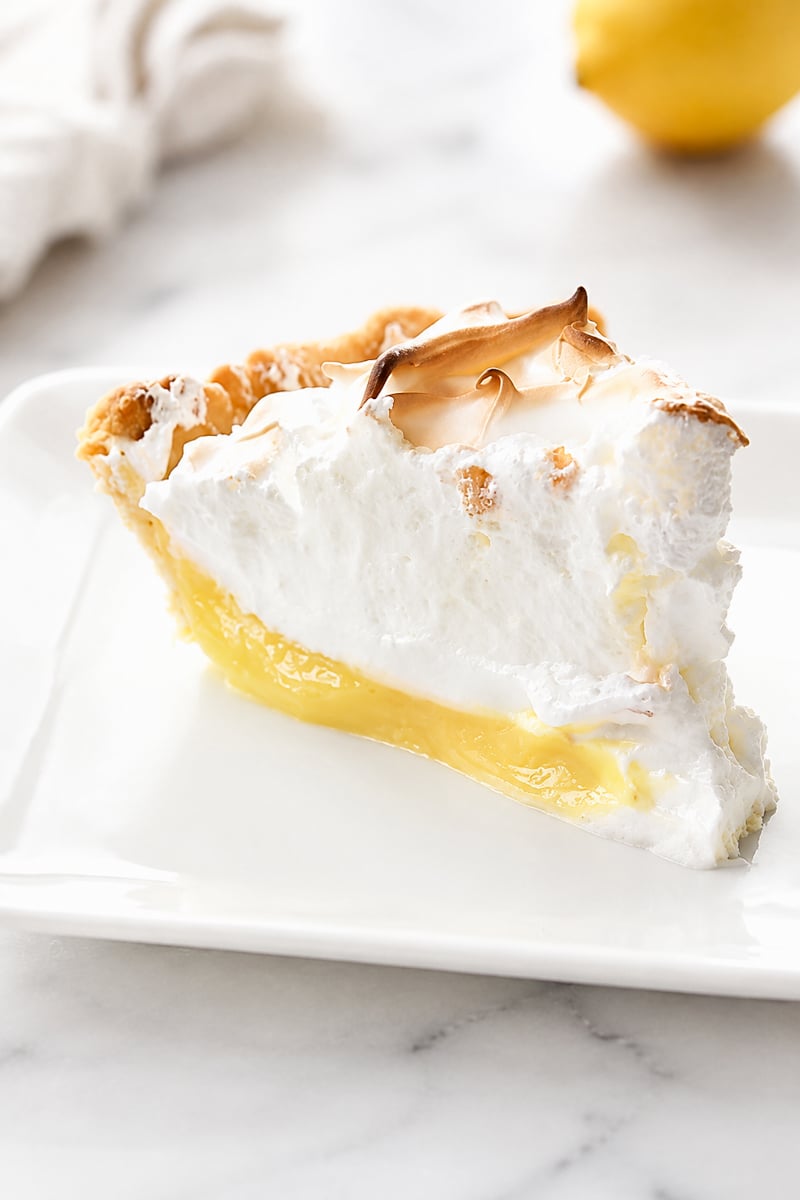 A slice of Lemon Meringue Pie with a golden-brown meringue topping sits on a white plate, with a lemon blurred in the background.
