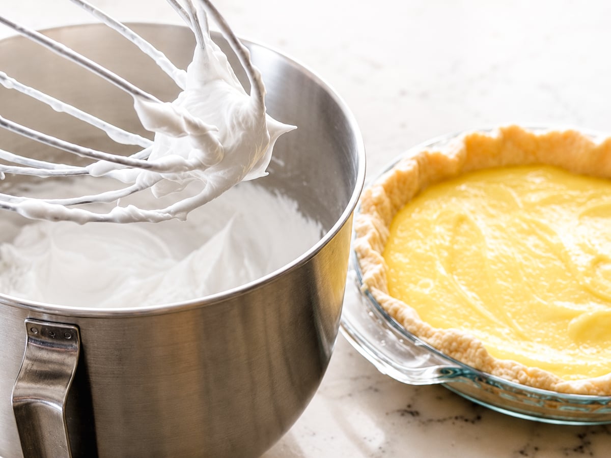 A metal bowl with whipped meringue and a whisk sits next to a glass pie dish filled with yellow custard and an unbaked pie crust on a light countertop, ready to be transformed into a classic Lemon Meringue Pie.