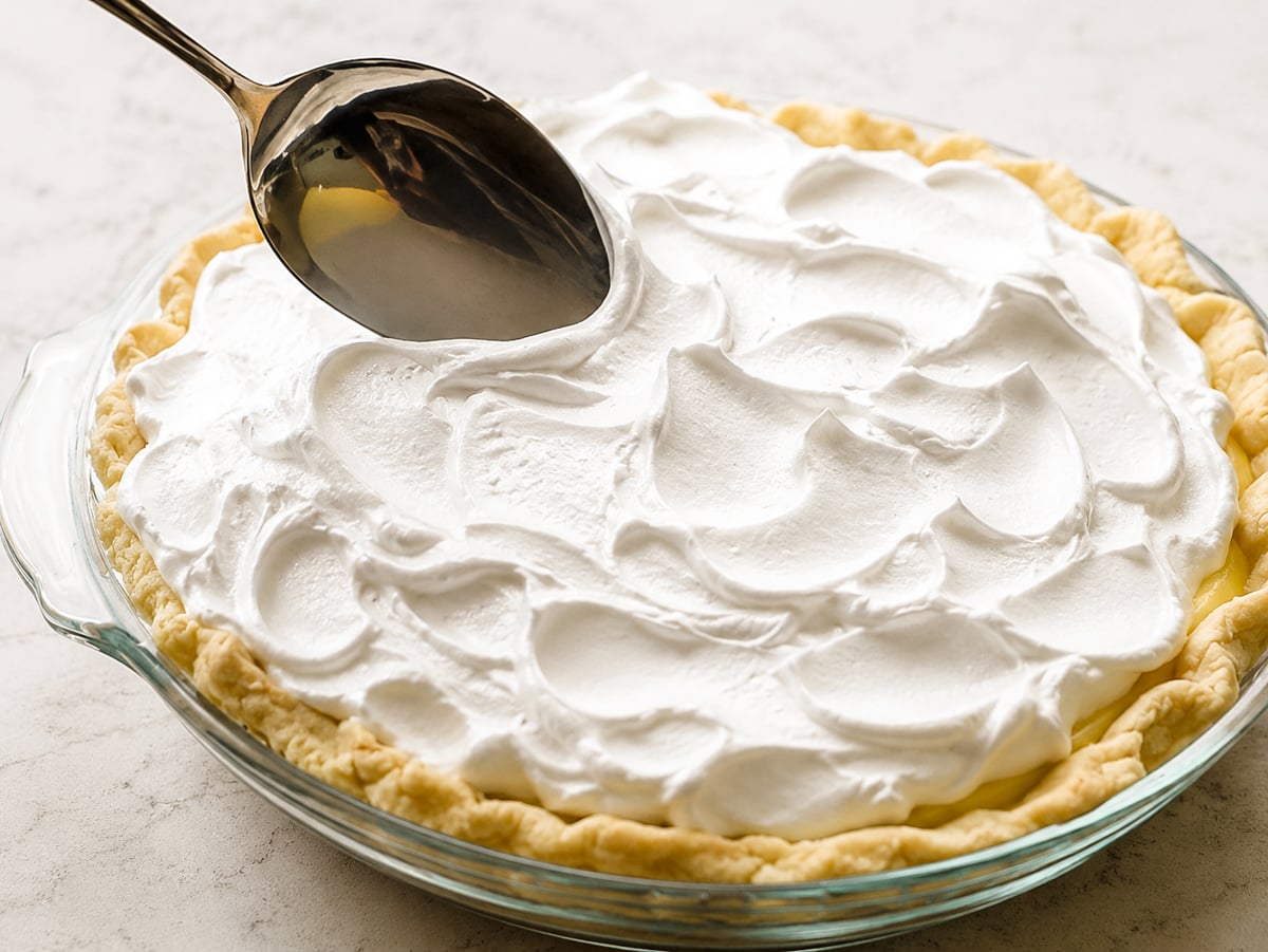 A spoon spreads fluffy white whipped topping over a Lemon Meringue Pie in a glass dish on a light countertop.