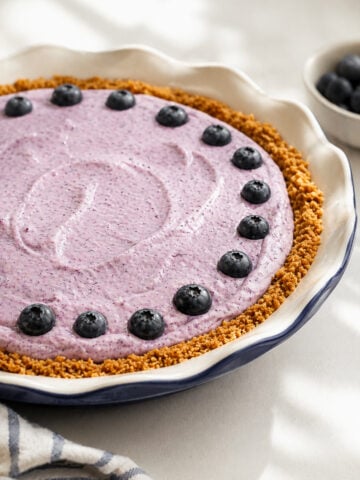 A Blueberry Icebox Pie in a scalloped dish features a golden graham cracker crust, luscious purple blueberry filling, and a ring of fresh blueberries. In the background, a bowl of extra blueberries completes the summery scene.