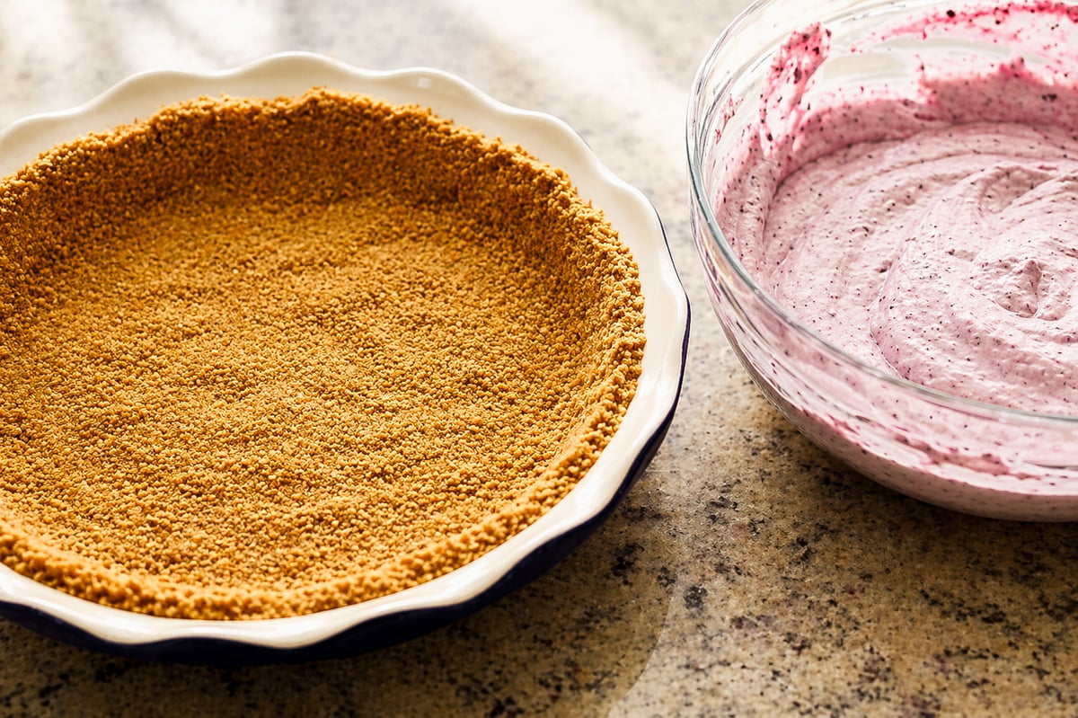 A graham cracker pie crust in a pie dish sits next to a glass bowl filled with a creamy, light purple blueberry icebox pie mixture on a speckled countertop.