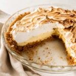 A partially eaten Key Lime Pie with a golden graham cracker crust, creamy yellow filling, and a fluffy, toasted meringue topping sits in a clear glass pie dish on a light surface with a beige cloth nearby.