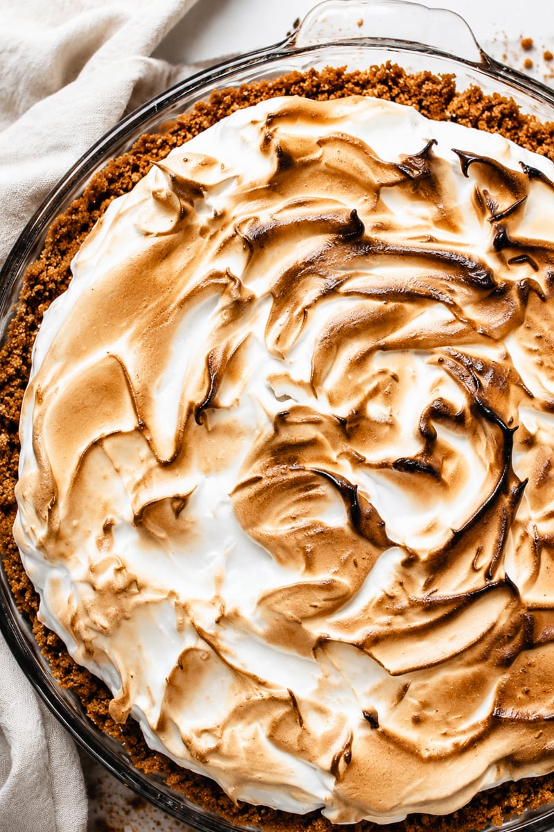 A close-up of a Key Lime Pie with a golden-brown, toasted meringue topping swirled on top of a crumbly crust in a glass pie dish, with a beige cloth partially visible beside it.