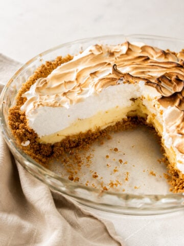 A partially eaten Key Lime Pie with a golden graham cracker crust, creamy yellow filling, and a fluffy, toasted meringue topping sits in a clear glass pie dish on a light surface with a beige cloth nearby.