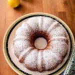 A Vegan Blueberry-Lemon Bundt Cake dusted with powdered sugar sits on a decorative plate, with two lemons and a patterned kitchen towel nearby on a wooden surface.