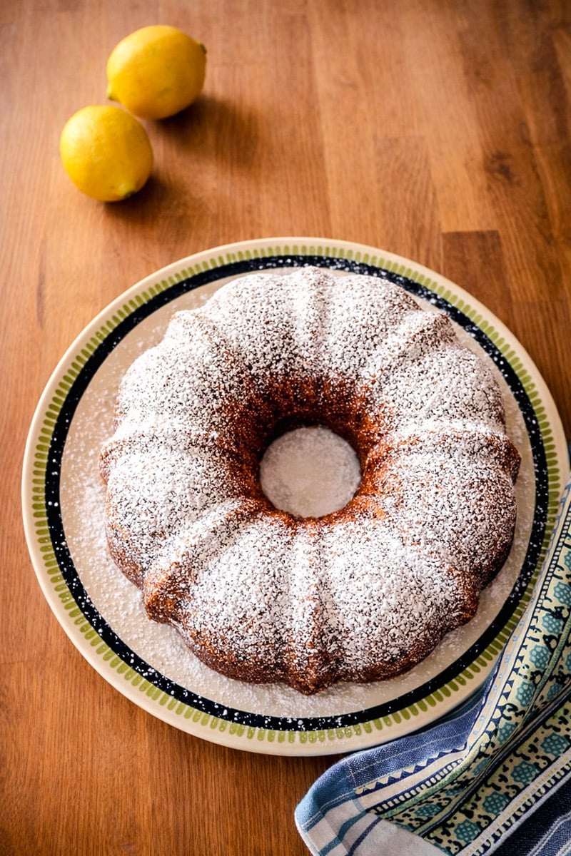 A Vegan Blueberry-Lemon Bundt Cake dusted with powdered sugar sits on a decorative plate, with two lemons and a patterned kitchen towel nearby on a wooden surface.
