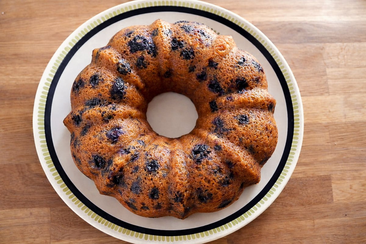 A golden-brown Vegan Blueberry-Lemon Bundt Cake with dark berry spots sits on a white plate with a patterned rim, placed on a wooden surface.