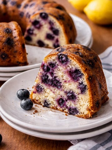 A slice of Vegan Blueberry-Lemon Bundt Cake on a white plate sits in front of the rest of the cake. Fresh blueberries and two lemons add a pop of color on the wooden surface, while a striped cloth is partially visible on the right.