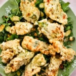 A green plate topped with several pieces of golden-brown, crispy Italian Stuffed Zucchini Flowers garnished with fresh greens and herbs, with small pieces of zucchini underneath.