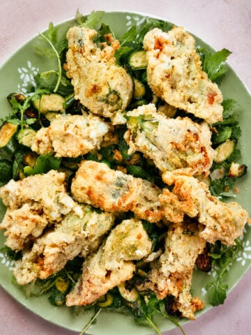 A green plate topped with several pieces of golden-brown, crispy Italian Stuffed Zucchini Flowers garnished with fresh greens and herbs, with small pieces of zucchini underneath.