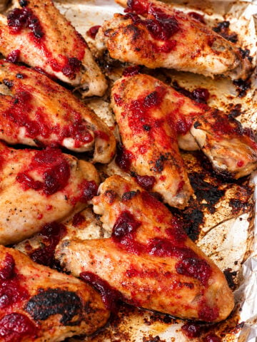 Baked chicken wings on a foil-lined tray, topped with a bright red cranberry sauce, with areas of slight charring and caramelization visible—reminiscent of the deep flavors found in slow roasted turkey wings.
