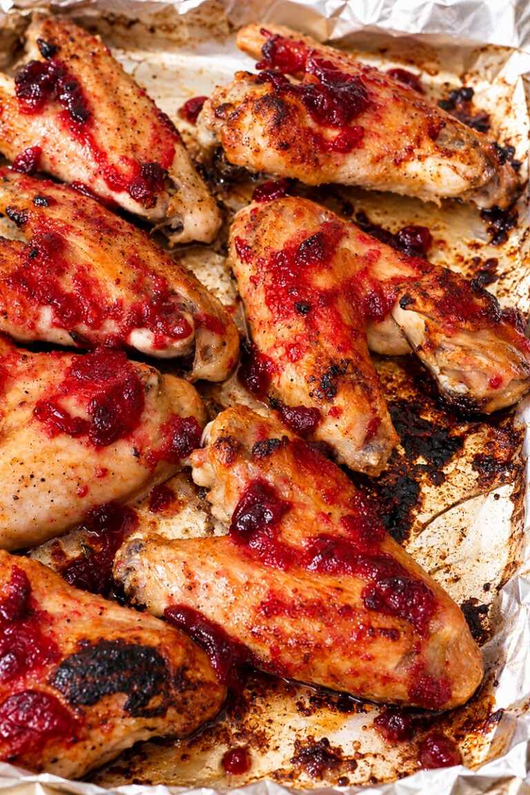Baked chicken wings on a foil-lined tray, topped with a bright red cranberry sauce, with areas of slight charring and caramelization visible—reminiscent of the deep flavors found in slow roasted turkey wings.