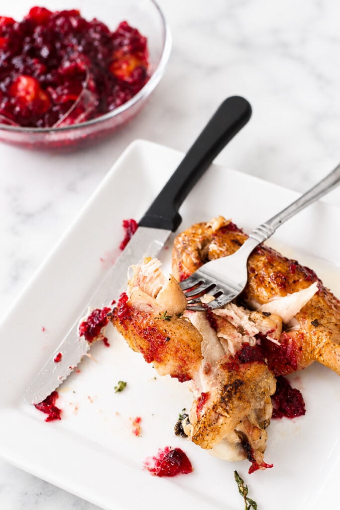 A plate with roasted chicken topped with cranberry sauce, a fork and knife cutting a piece, and a bowl of cranberry sauce in the background on a marble surface—perfectly capturing the cozy comfort of slow roasted turkey wings served during holiday gatherings.