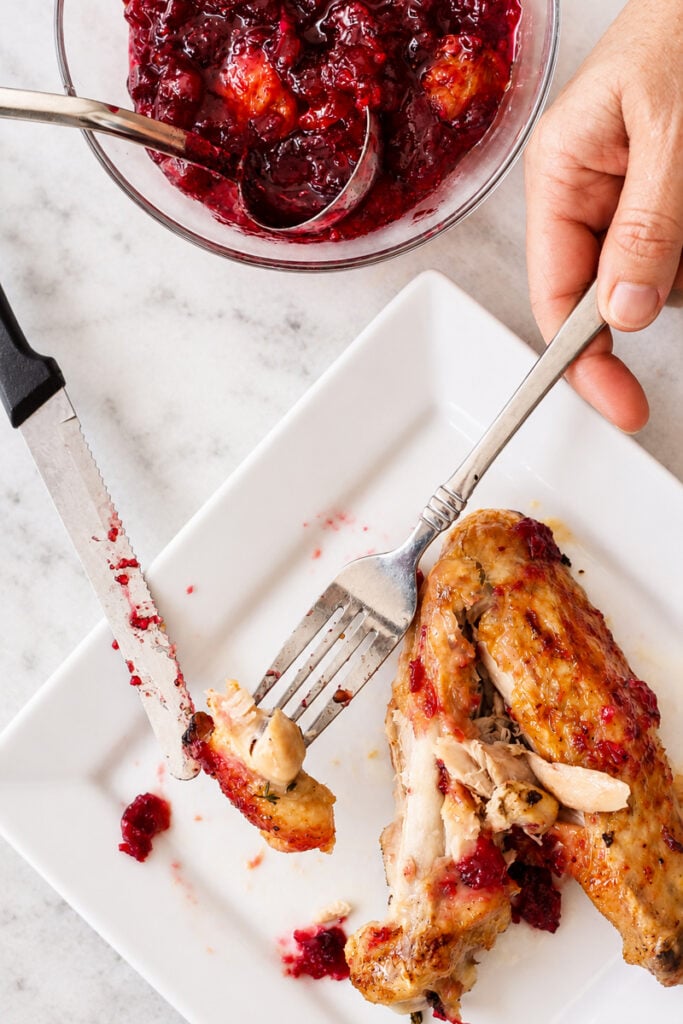 A hand uses a fork and knife to cut into slow roasted turkey wings topped with cranberry sauce on a white plate, with a bowl of cranberry sauce nearby.
