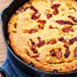 Savory cornbread topped with crispy bacon pieces, baked to a golden brown, sits on a blue cloth on a wooden table.