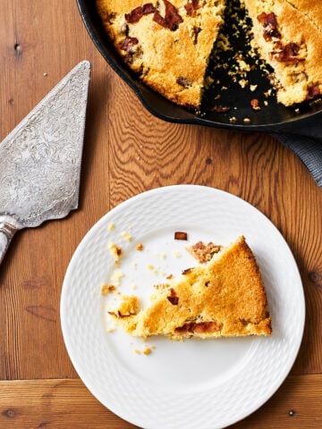 A slice of savory cornbread with bacon sits on a white plate, with crumbs scattered. The rest of the cornbread is in a skillet nearby, and an ornate pie server rests on the wooden table.
