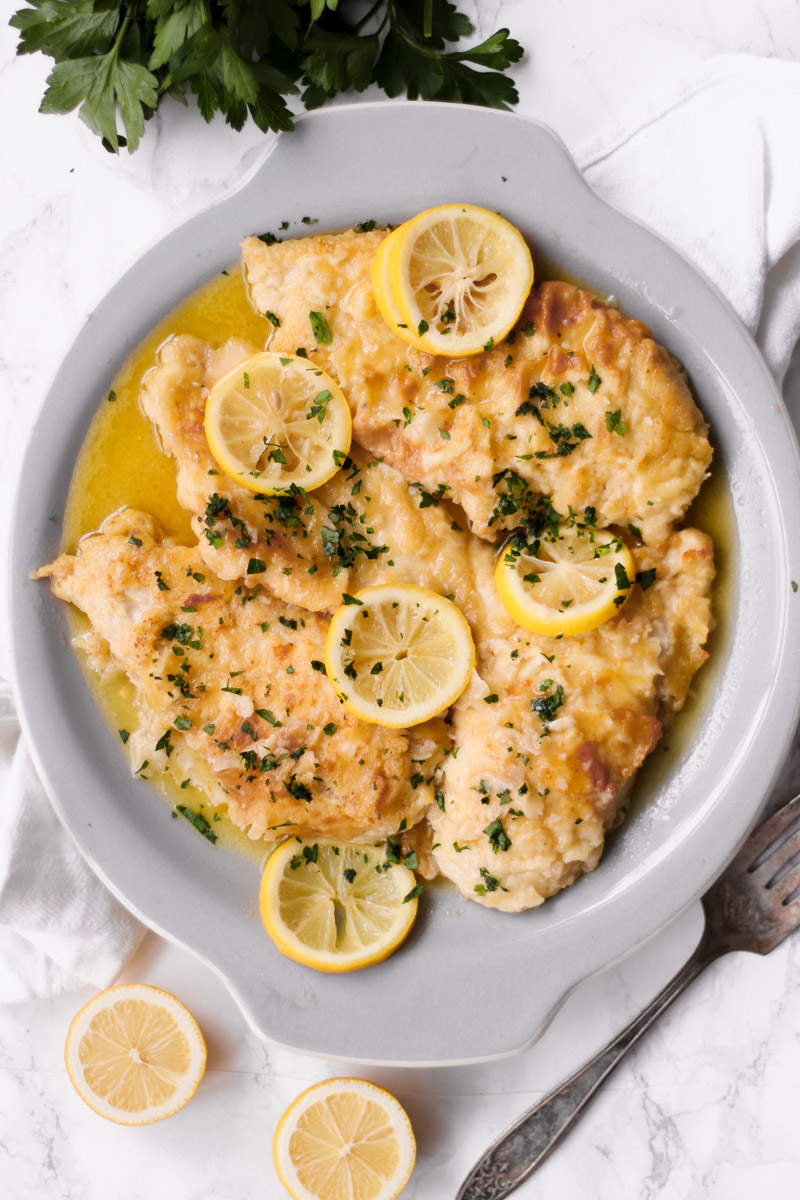 image of cooked chicken slices with lemon on a serving plate with fork in background.