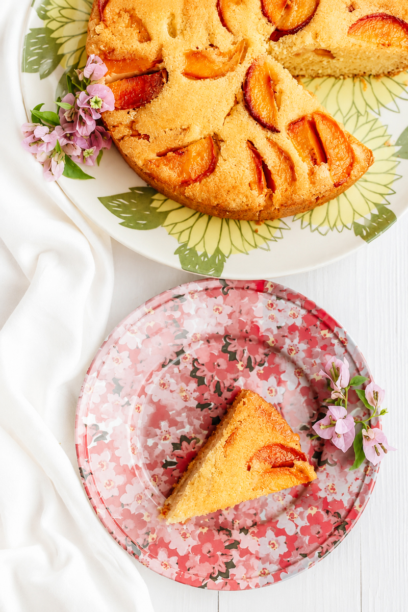A slice of vegan peach polenta cake on a floral plate sits beside the remaining cake on a green patterned platter. Both plates are decorated with small purple flowers and set on a white tablecloth.