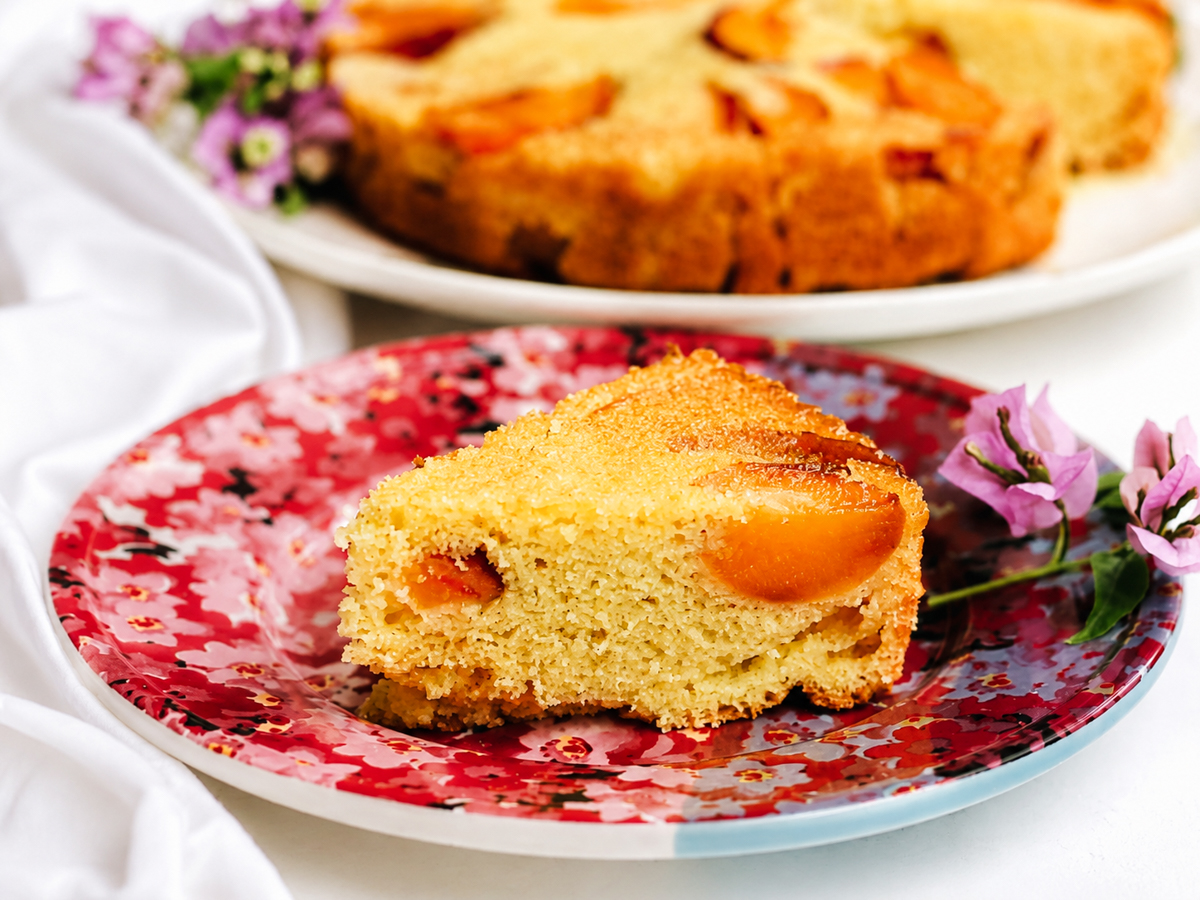 A slice of golden vegan peach polenta cake with peach pieces sits on a red floral plate, with a whole cake and purple flowers visible in the background.