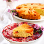 A slice of vegan peach polenta cake on a floral-patterned plate, garnished with purple flowers. The remaining cake and a pot of pink flowers are blurred in the background.