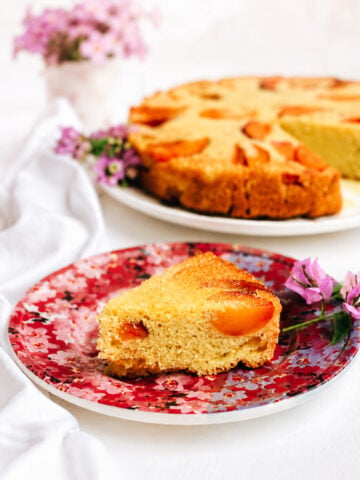 A slice of vegan peach polenta cake on a floral-patterned plate, garnished with purple flowers. The remaining cake and a pot of pink flowers are blurred in the background.