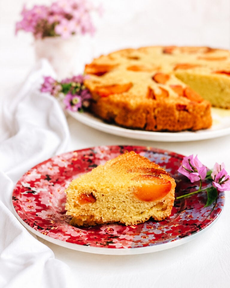A slice of vegan peach polenta cake on a floral-patterned plate, garnished with purple flowers. The remaining cake and a pot of pink flowers are blurred in the background.