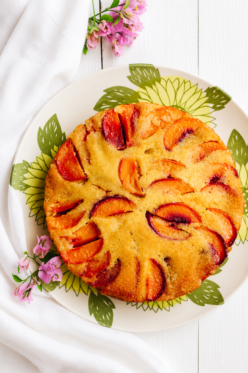 A vegan peach polenta cake with a golden brown crust and sliced plums on top, arranged on a plate with green leaf patterns, accented by pink flowers and a white cloth on a white wooden surface.