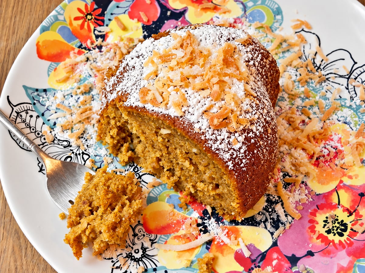 A slice of golden pumpkin cake topped with powdered sugar and toasted coconut on a floral plate, with a fork holding a small piece of the cake.